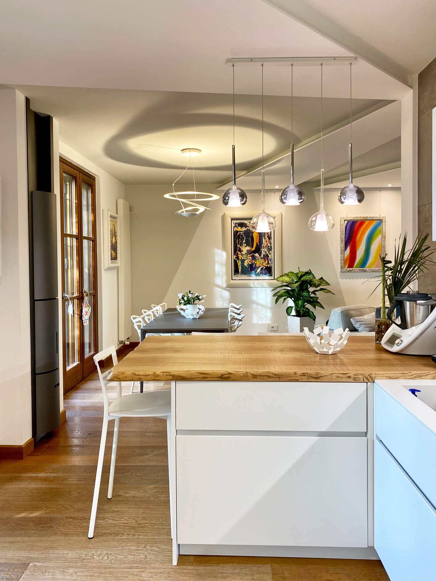 La zona giorno vista dalla cucina: il piano in legno massello, le piante, le lampade a sospensione e il tavolo da pranzo sullo sfondo